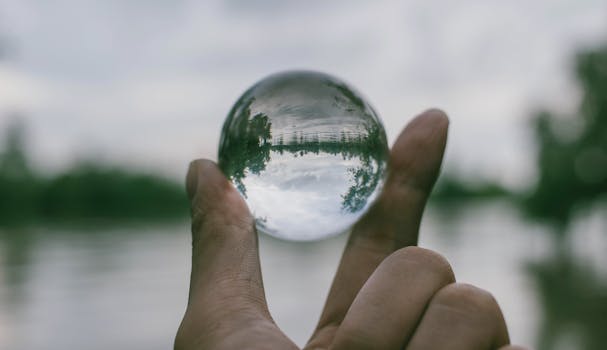 A hand holding a transparent glass sphere outdoors, showcasing an inverted reflection of the landscape.